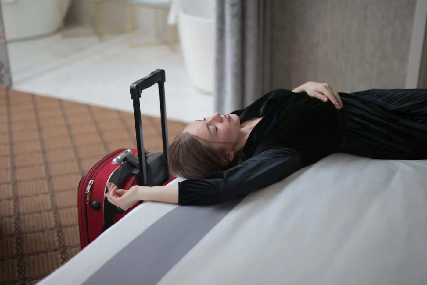 An exhausted traveler resting on a hotel bed beside her suitcase