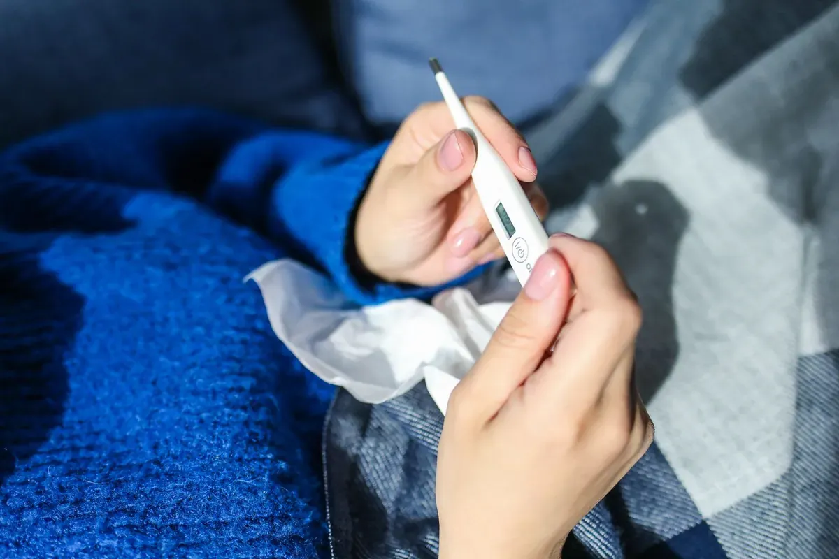 A person holding a thermometer while resting under a blanket